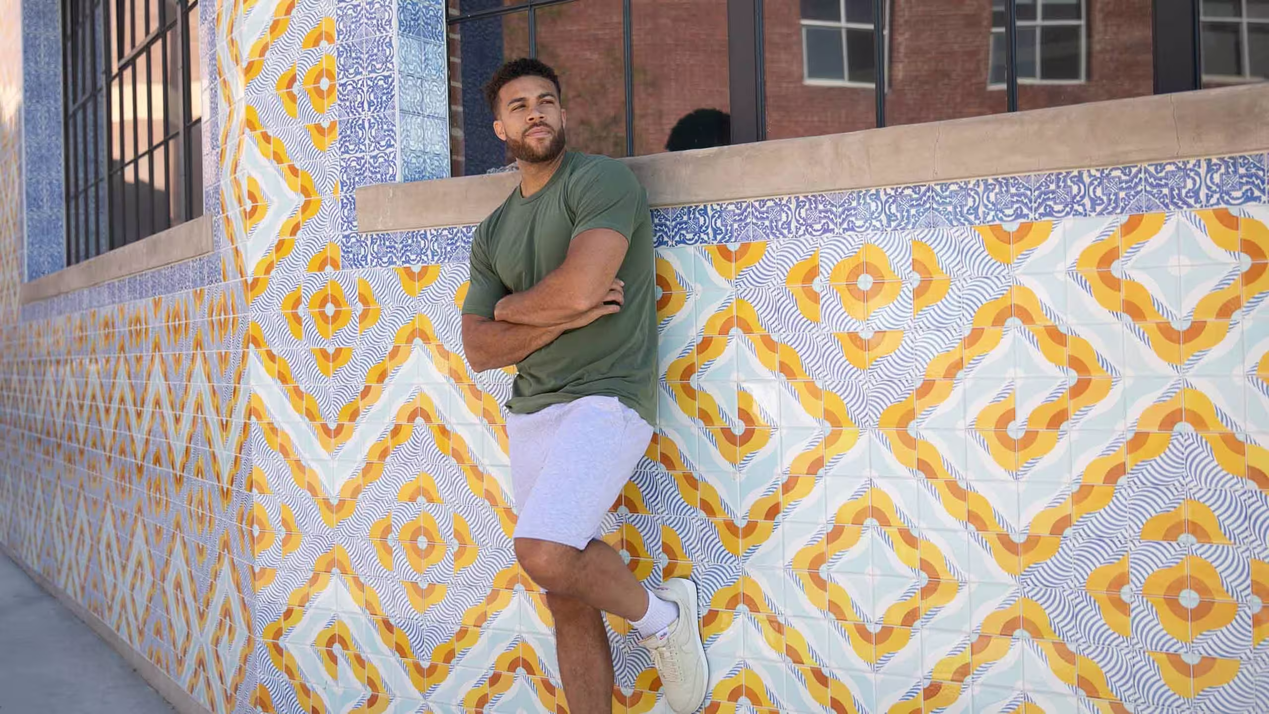 man wearing a dark green t-shirt and pink shorts leaning against a wall