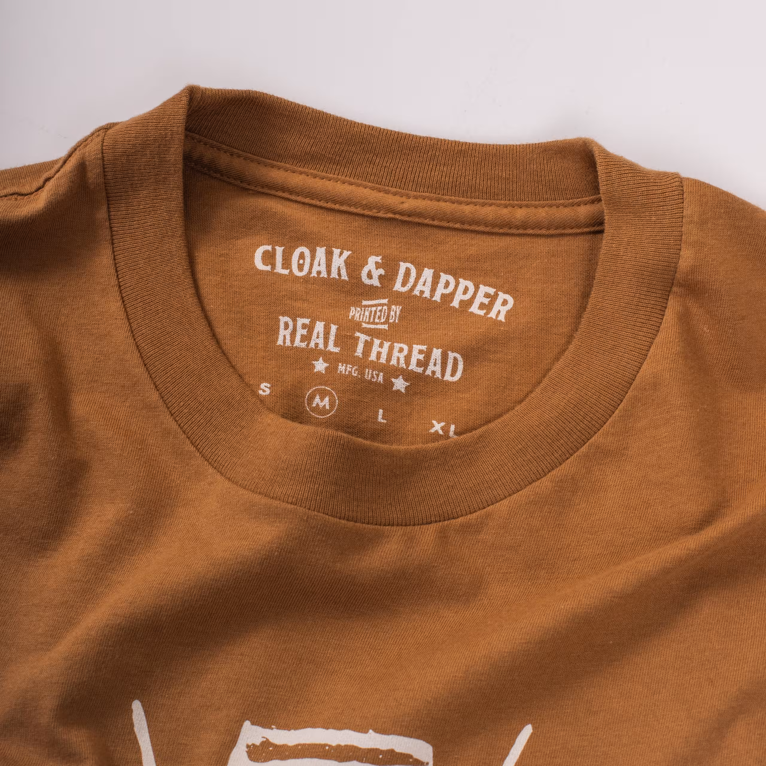 A burnt orange shirt with a custom printed tag that says "cloak and dapper, printed by Real Thread" 