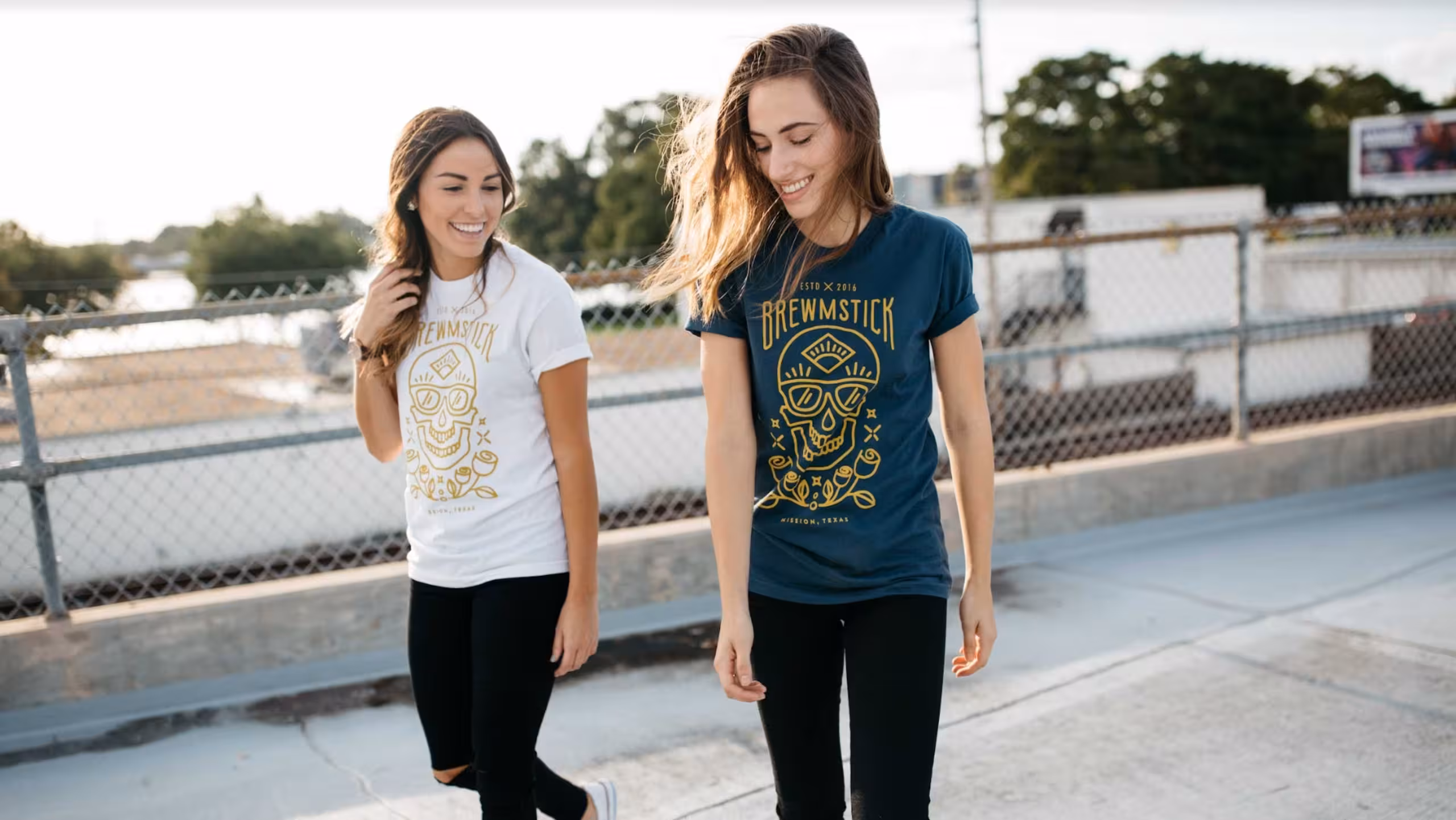 Two women walking and smiling on a rooftop, wearing black pants and graphic t-shirts, one white and one blue, with a chain-link fence in the background.