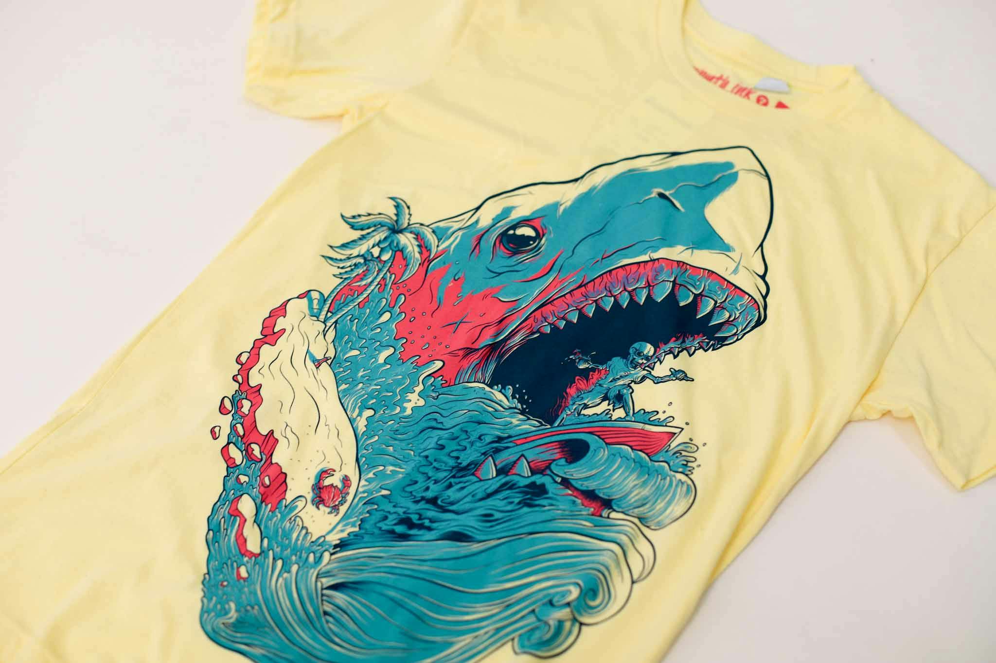 Yellow shirt with artwork of blue and pink shark with skeleton surfing in its mouth Yellow shirt with artwork of blue and pink shark with skeleton surfing in its mouth